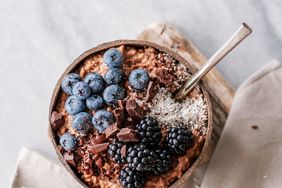 A bowl of oatmeal topped with blueberries blackberries shredded coconut and chocolate chunks on a table with a napkin