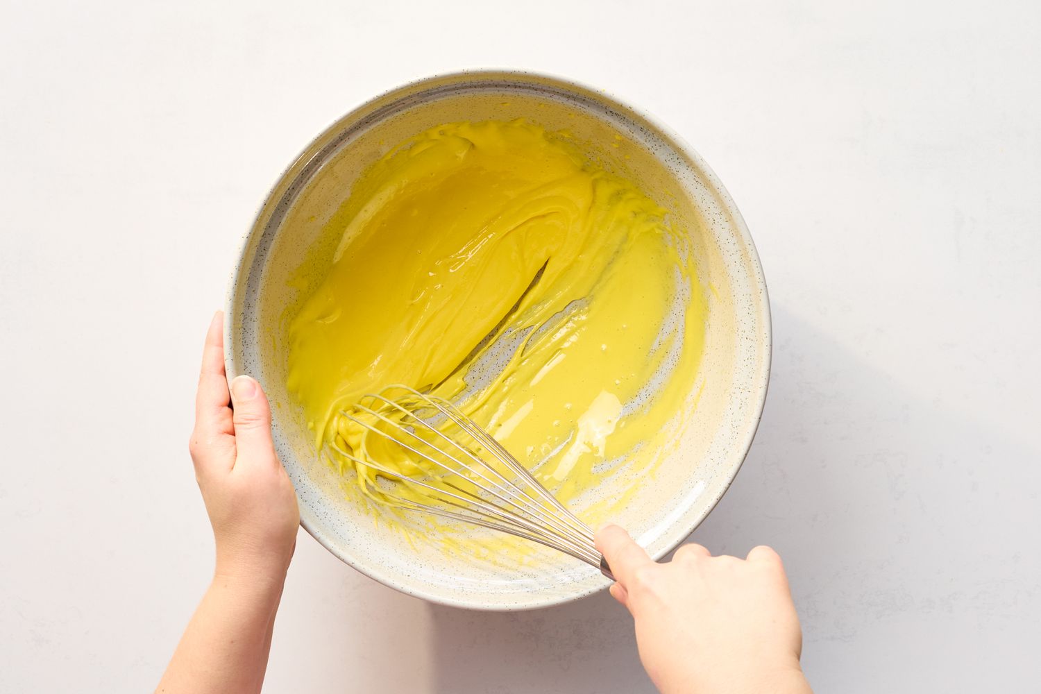 Hands holding a bowl with a whisk stirring a yellow mixture indicative of preparation for making eggnog or a similar recipe