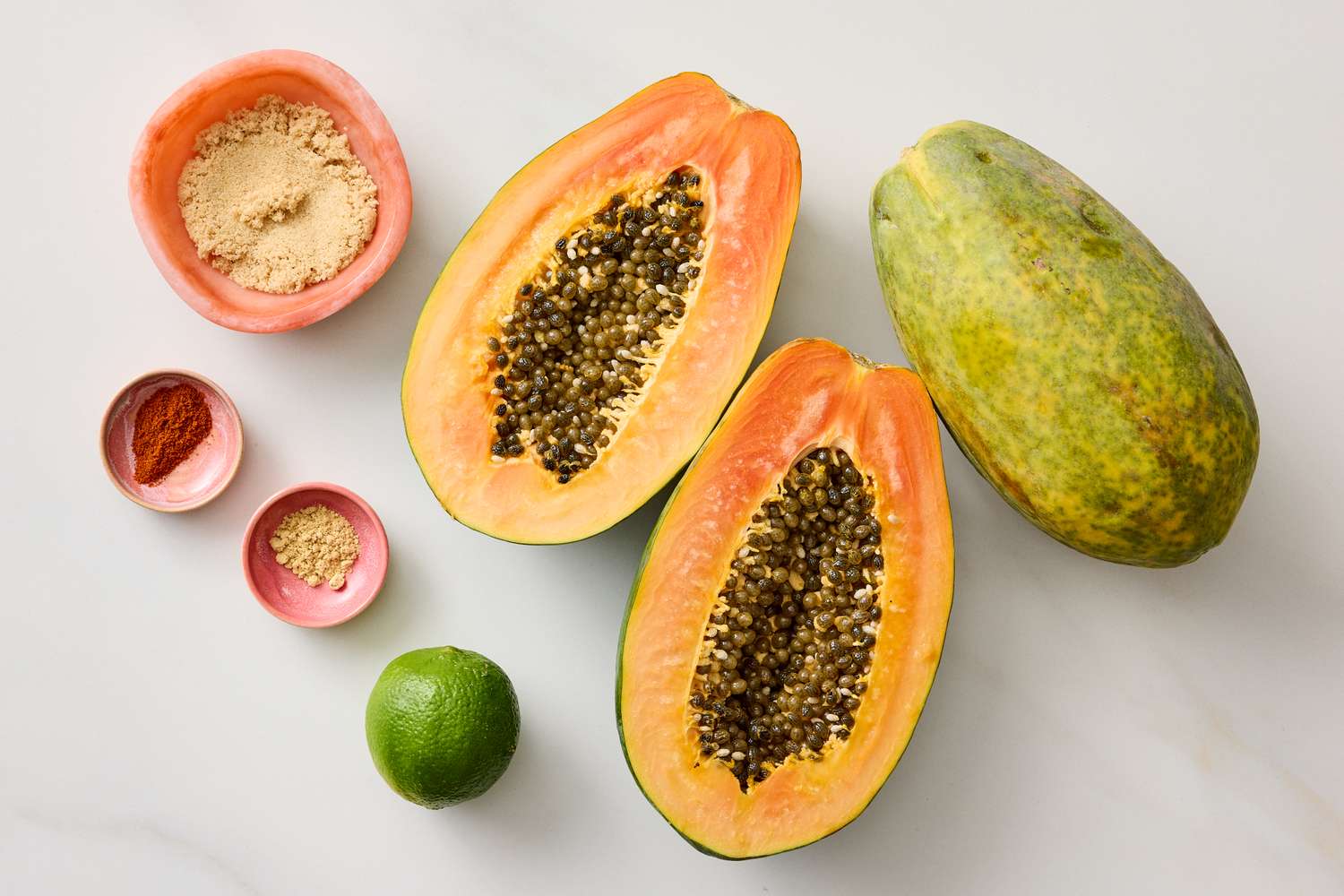 Sliced papaya with various small bowls of spices and a lime on a white surface
