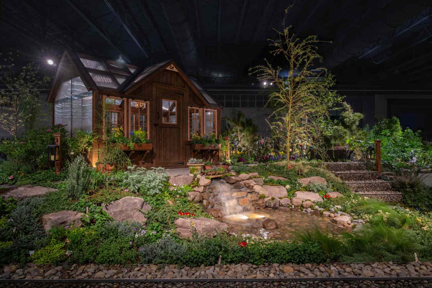 A landscaped garden with a wooden shed plants a water feature and natural paths in an indoor exhibit setting