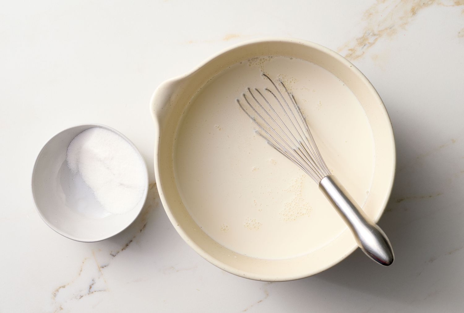 A bowl with a whisk placed inside and a smaller empty bowl next to it, both on a marble countertop