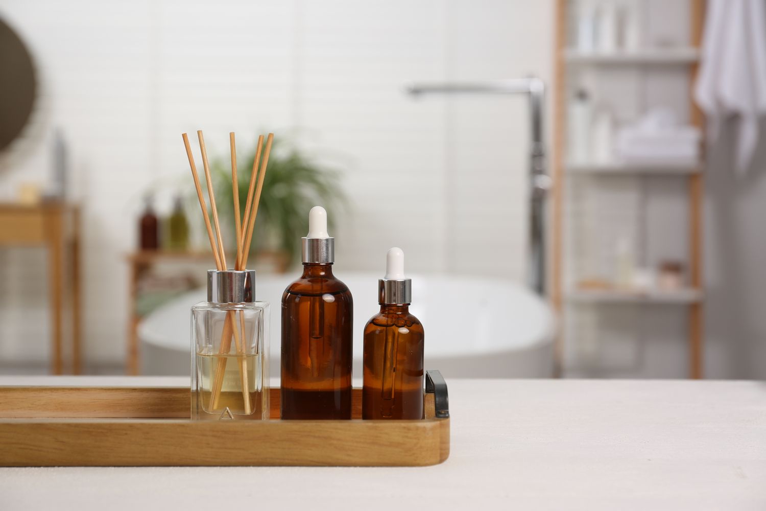 Three amber glass bottles and a reed diffuser on a wooden tray with a blurred bathroom background featuring a bath and shelves