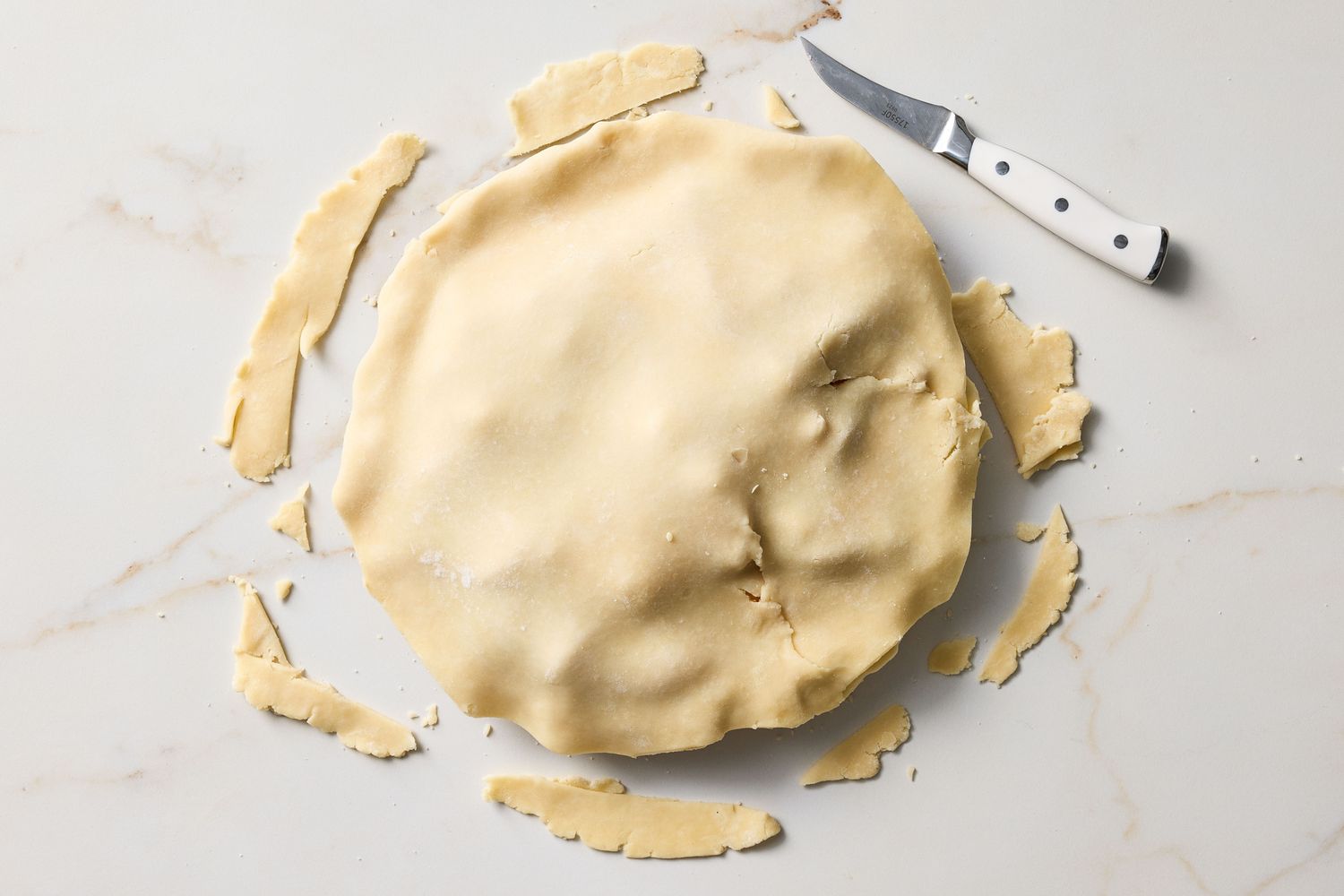 Unbaked pie with trimmed dough pieces and a knife on a countertop
