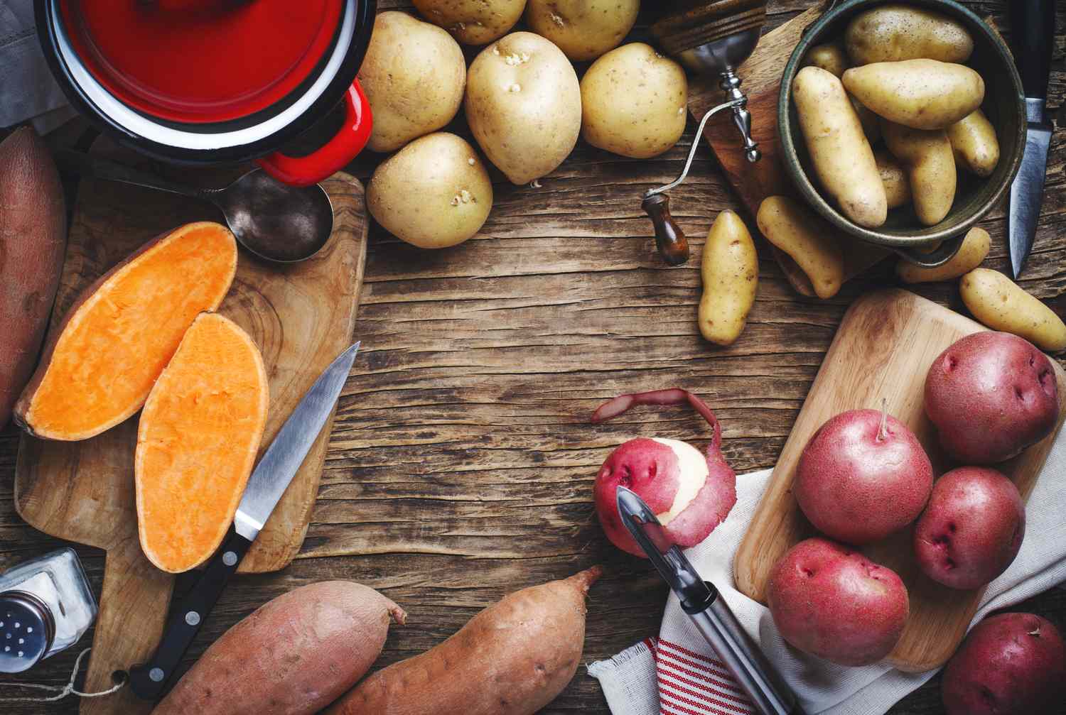 raw sweet potatoes and 3 types of white potatoes on wood surface with peeler and knives