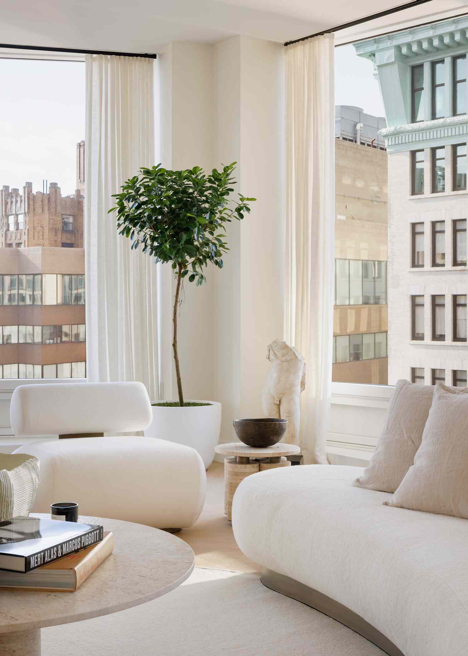 Minimalist living room by French California: white sculptural sofa and chairs, potted ficus tree, classical bust, and neutral decor. Large windows with sheer curtains offer city views, creating a calm, elegant space bathed in natural light.