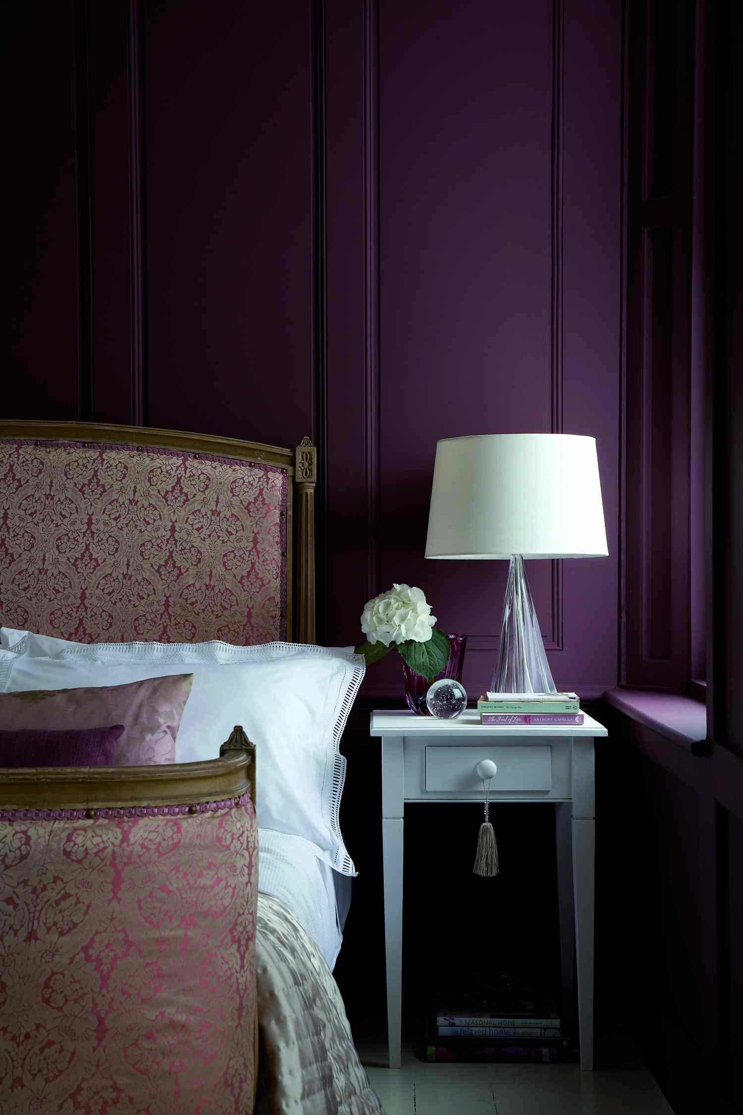 A bedroom with an ornate bed a nightstand holding a lamp and flower vase adjacent to a paneled wall