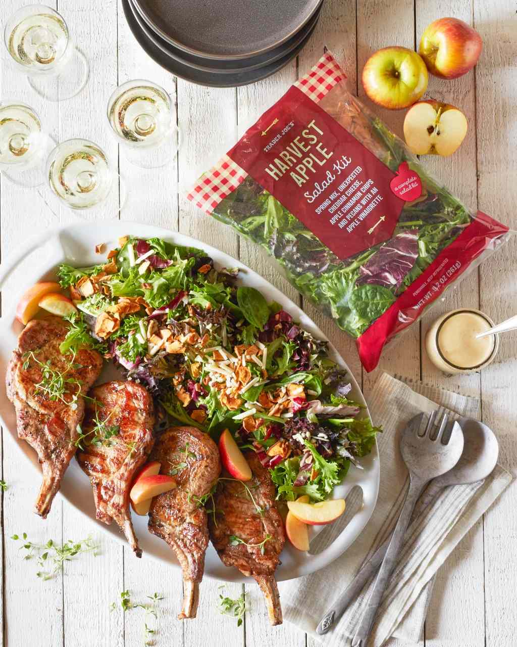 An arranged meal featuring pork chops a salad with apple slices and a packaged Harvest Apple salad kit on a table