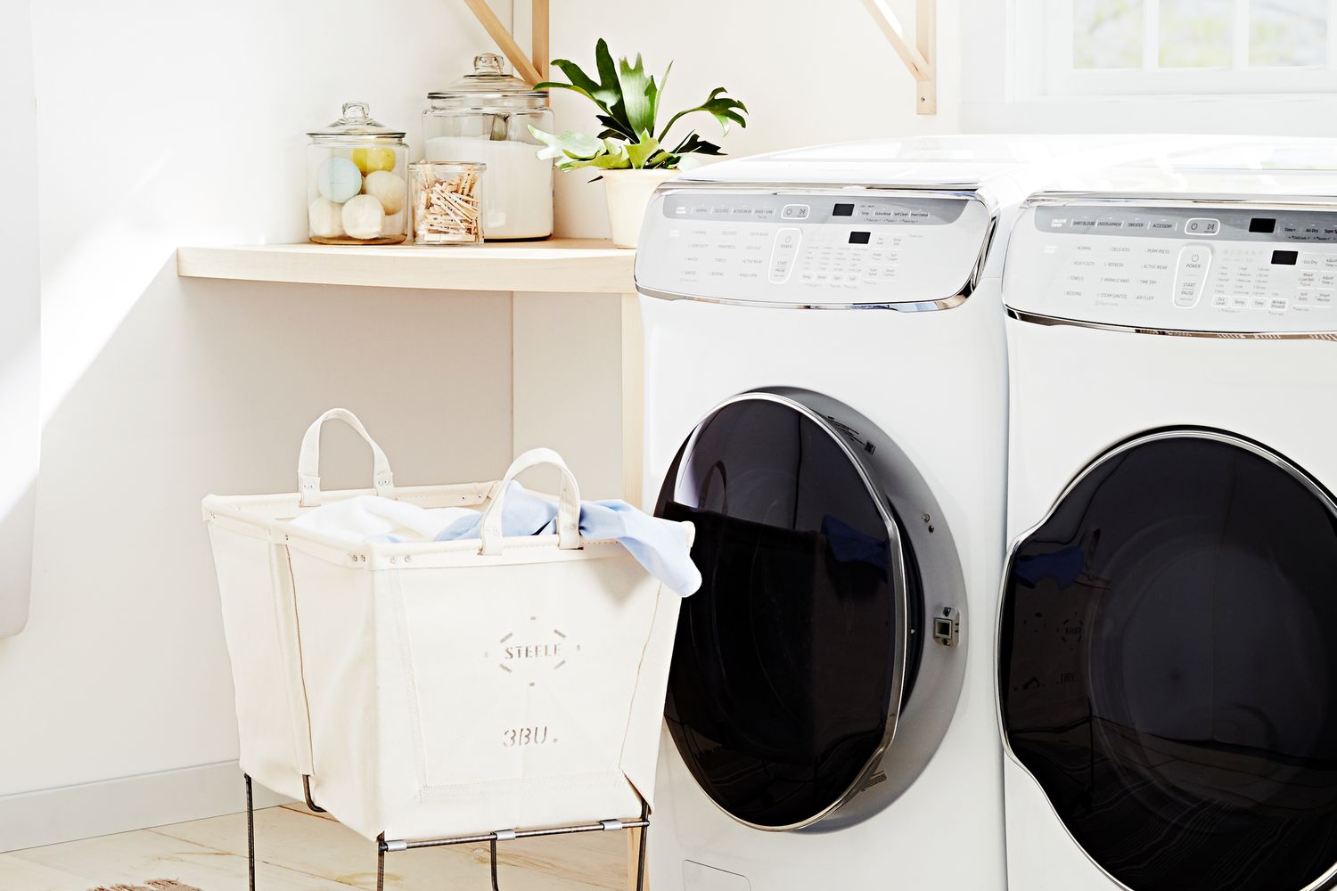 A laundry room setup featuring a washer and dryer beside a cart with laundry bags with shelves and decorations in the background
