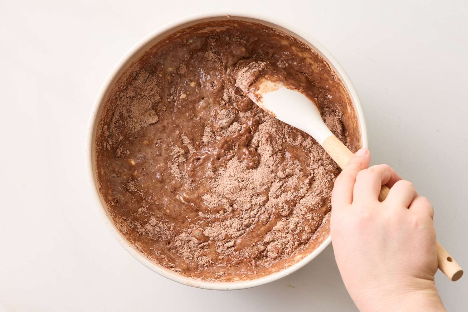 Bowl of chocolate cupcake batter being mixed with a spatula by a persons hand