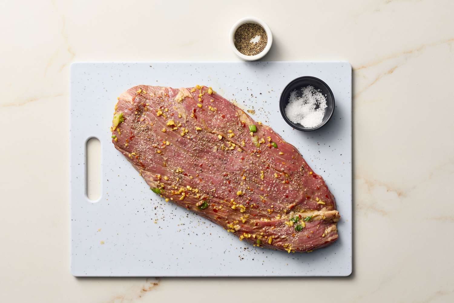 Flank steak marinated with garnishes on a cutting board, salt and pepper in small bowls nearby