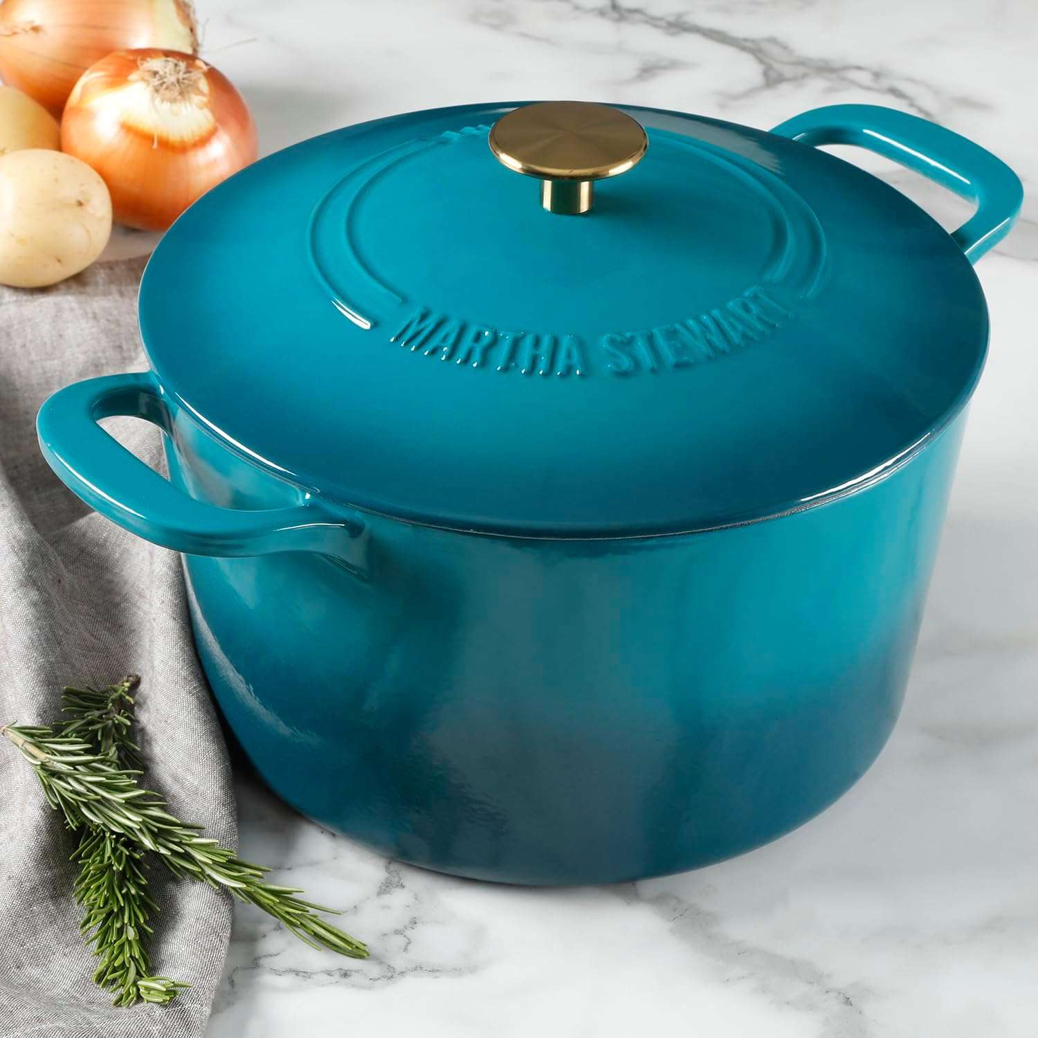 A cast iron cookware with a lid and handles with the brand name embossed on top placed on a countertop with aromatic vegetables nearby