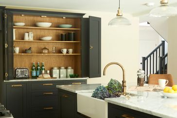A kitchen with a farmhouse sink, dark cabinetry, open shelving with dinnerware, and brass fixtures