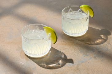 Two gin gimlet cocktails garnished with lime wedges on a surface with light and shadow patterns