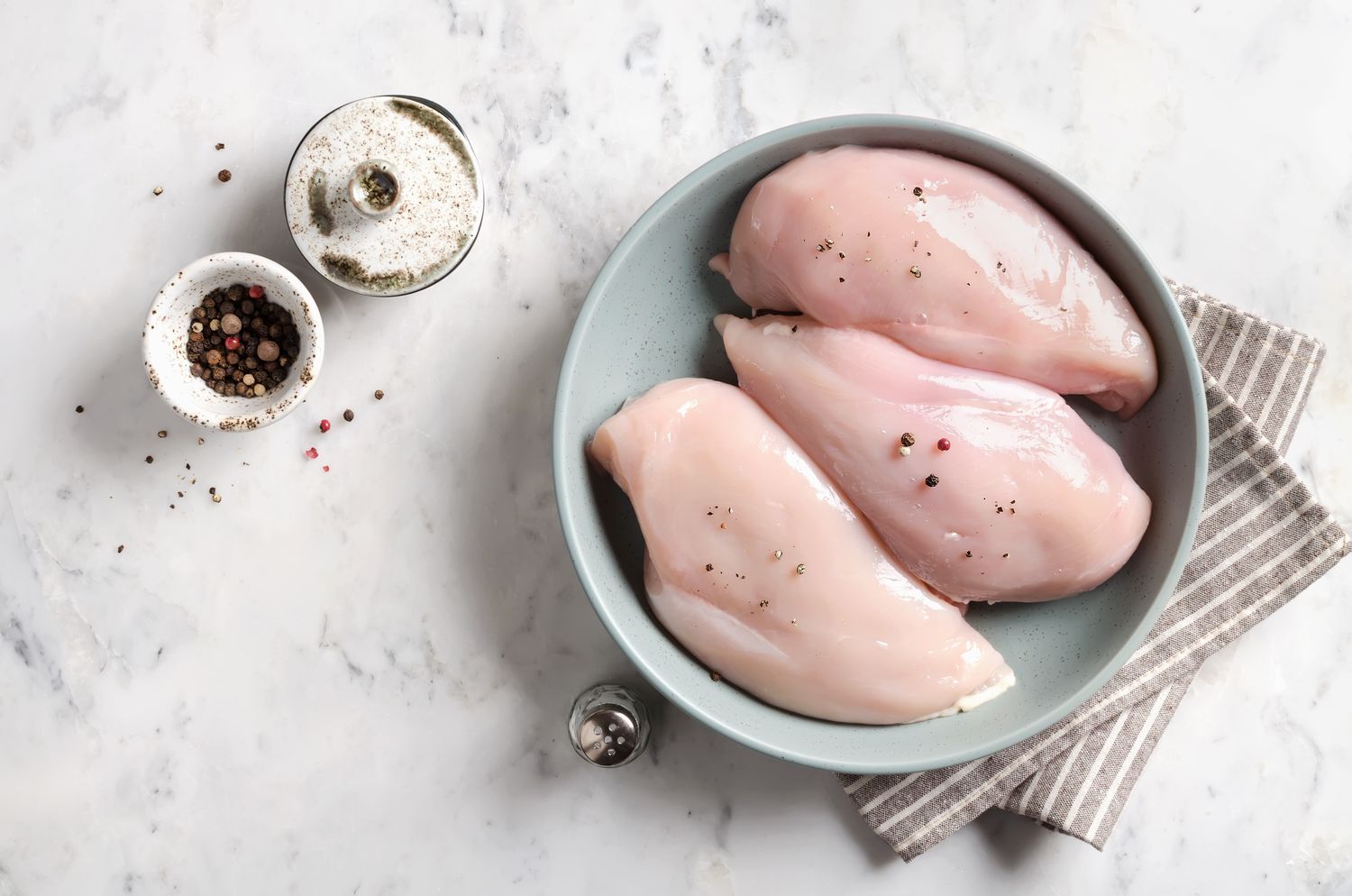 Raw chicken breasts in a bowl with seasoning ingredients nearby including salt and pepper on a marble surface