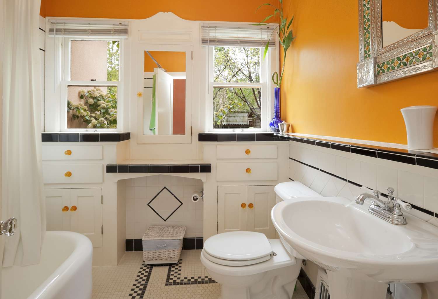 A bathroom with a pedestal sink a bathtub and orange walls featuring black and white tiled decor