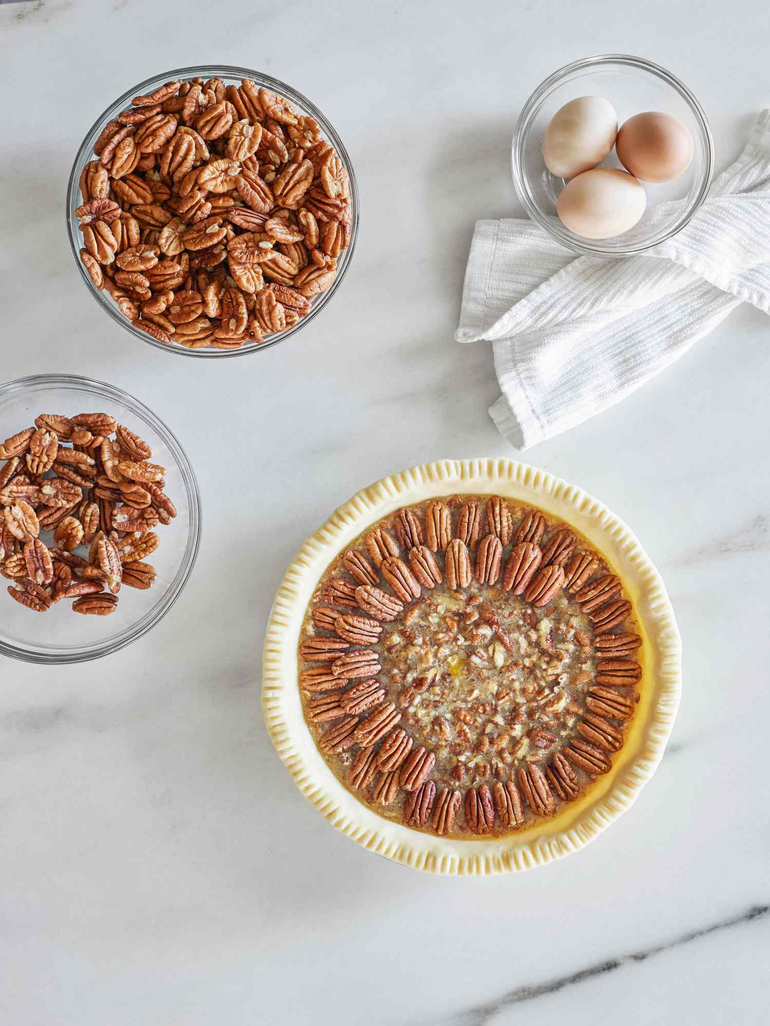 Pecan pie with bowls of pecans and eggs on a white surface