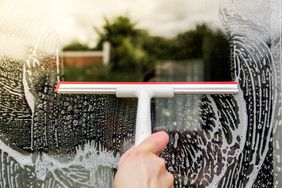 A hand using a squeegee to clean a soapy window outdoor view visible through the glass
