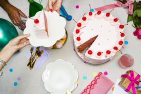 A festive table with a pink frosted cake a slice being served surrounded by plates forks balloons and party decorations