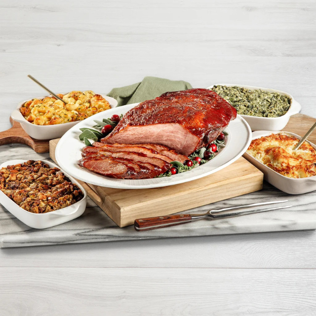 A cooked ham on a platter surrounded by various side dishes on a marble and wood serving board