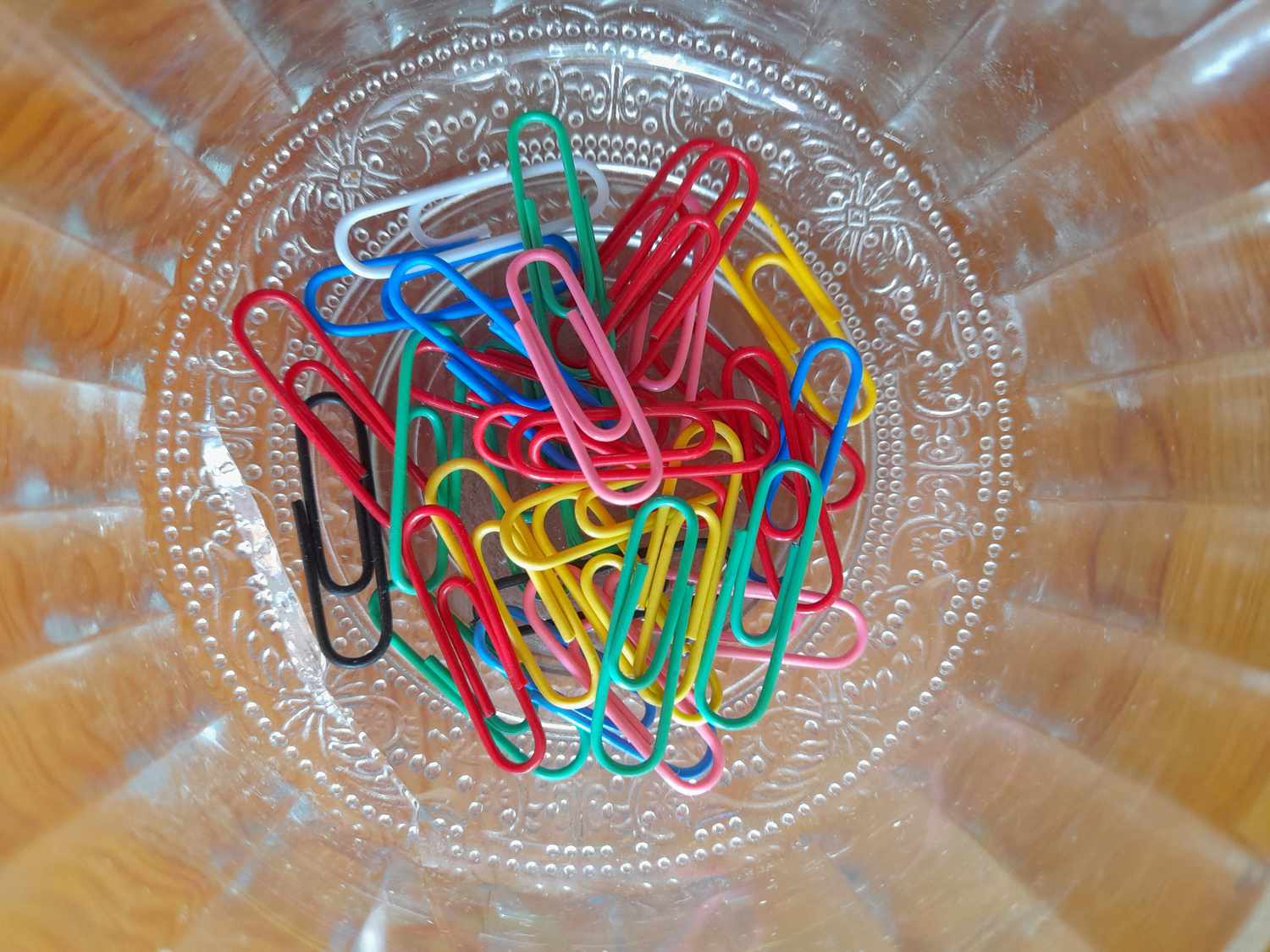 A collection of paperclips in various colors resting within a glass container viewed from above