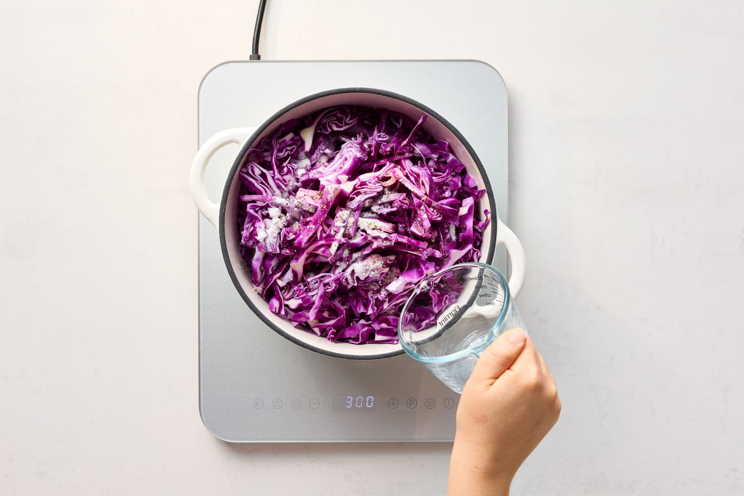 A pot of red cabbage and onions being cooked on a stove with a hand pouring liquid from a cup into the pot