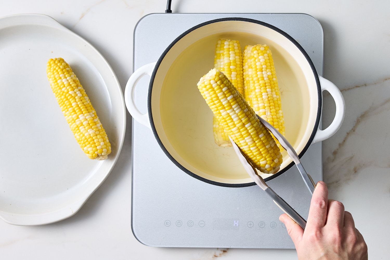 Corn cobs being boiled in a pot on an induction stovetop using tongs with a plate containing cooked corn in view