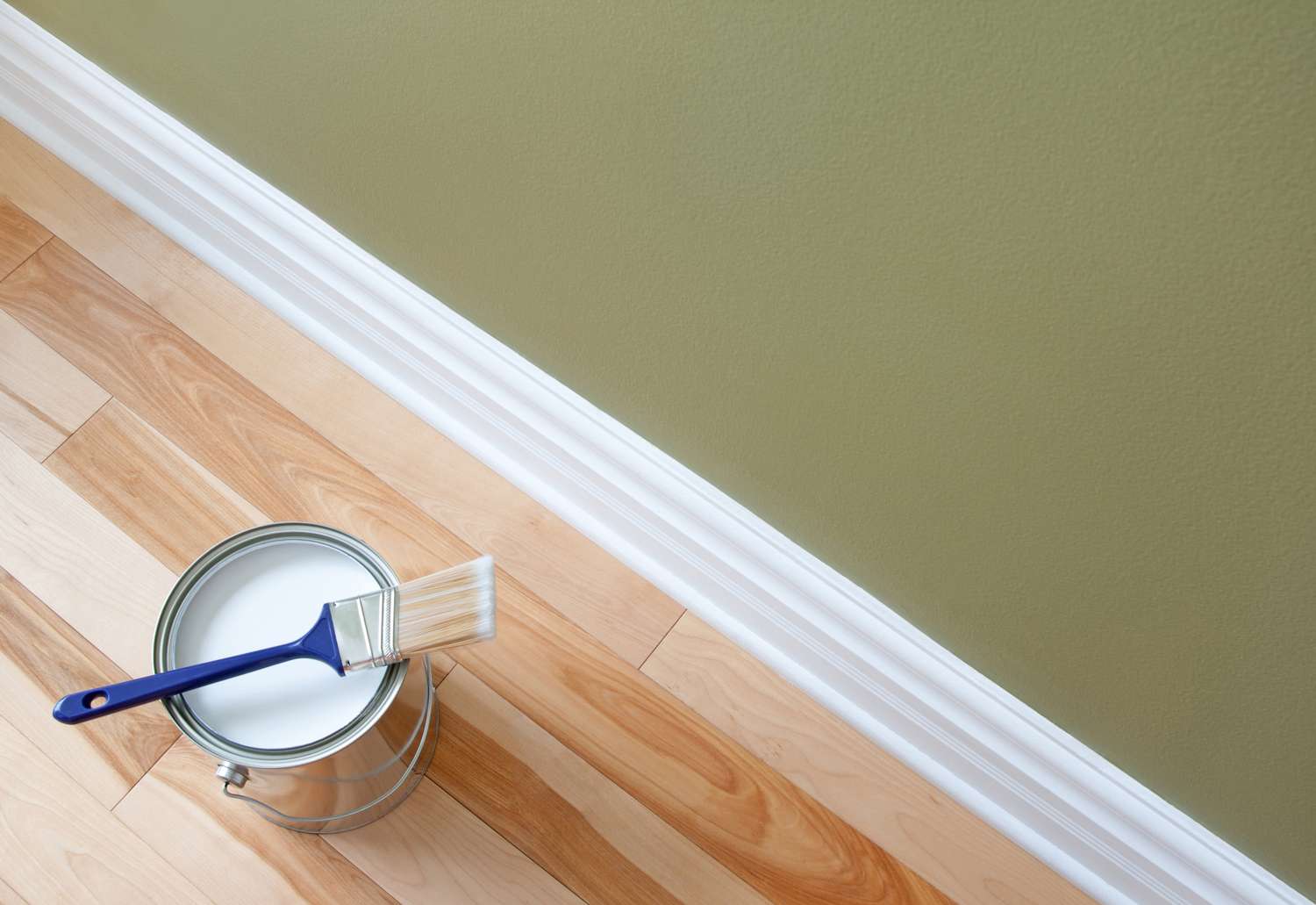 Can of paint with a brush placed on hardwood floor near a baseboard partially painted wall in view