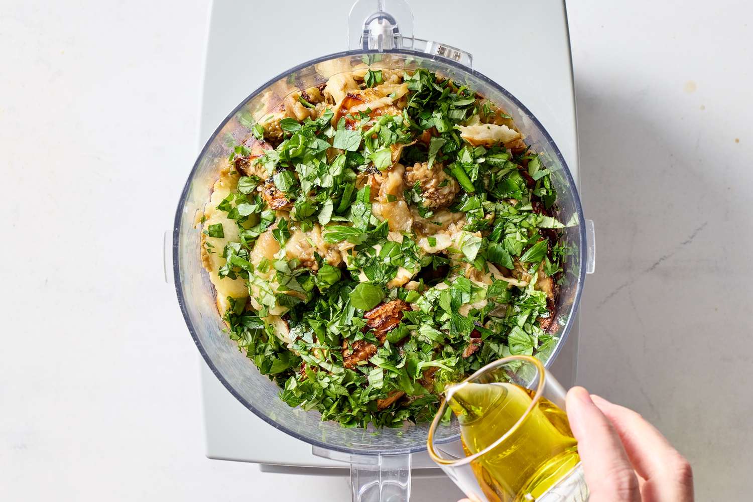 A food processor containing ingredients being blended for a dip recipe with a hand pouring olive oil into it