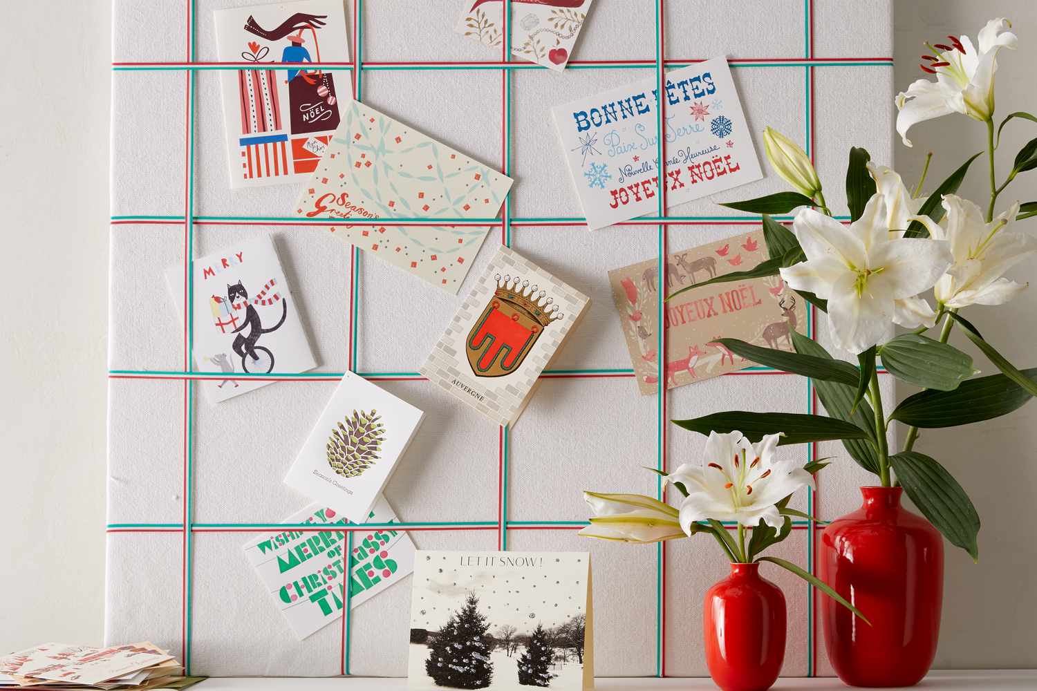 A display of Christmas cards pinned to a grid accompanied by white lilies in red vases on a table