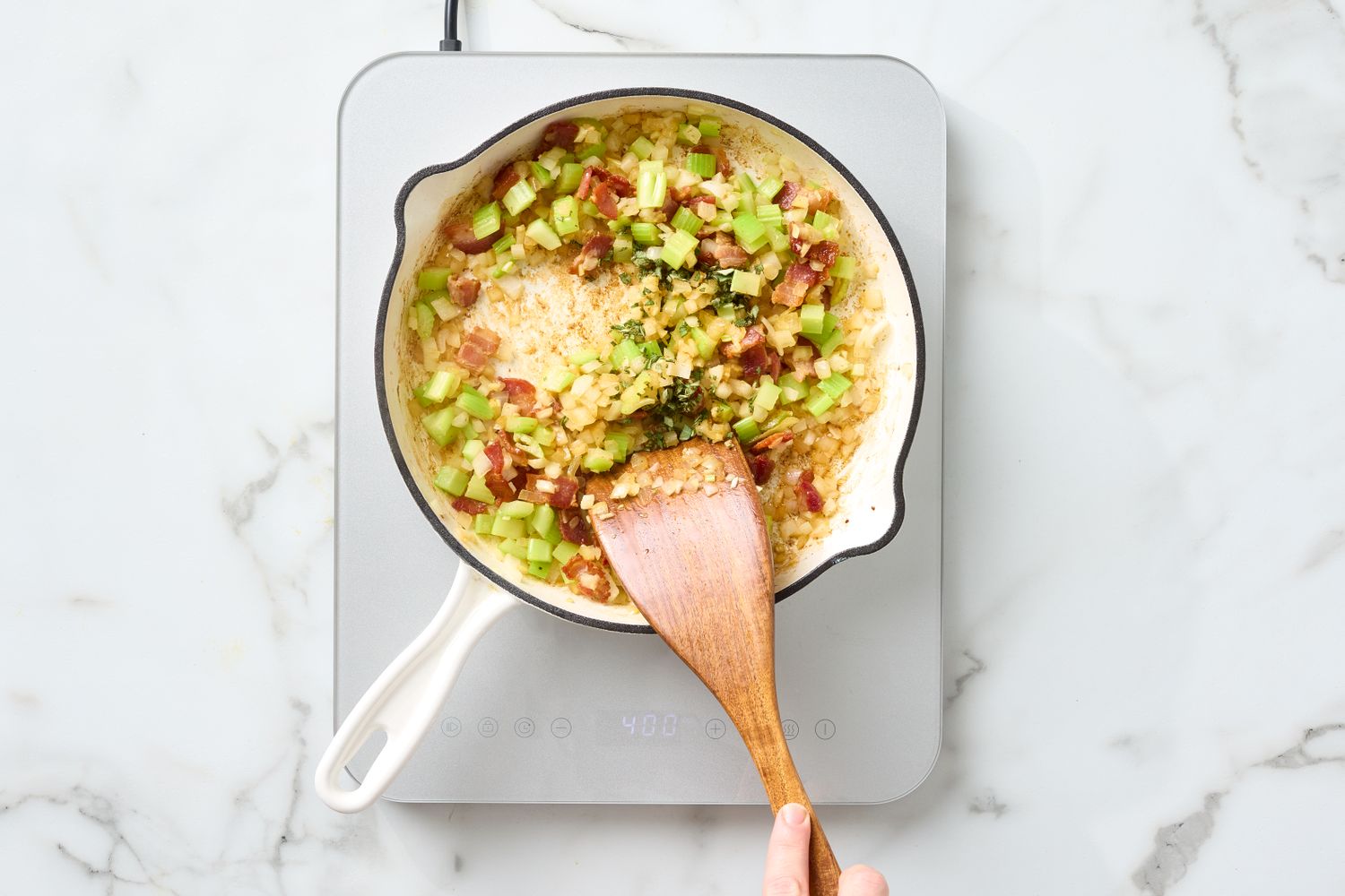 A skillet on a hot plate containing ingredients like celery bacon and cornbread being mixed with a wooden spatula