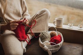 A person knitting by a window with a basket containing skeins of yarn nearby