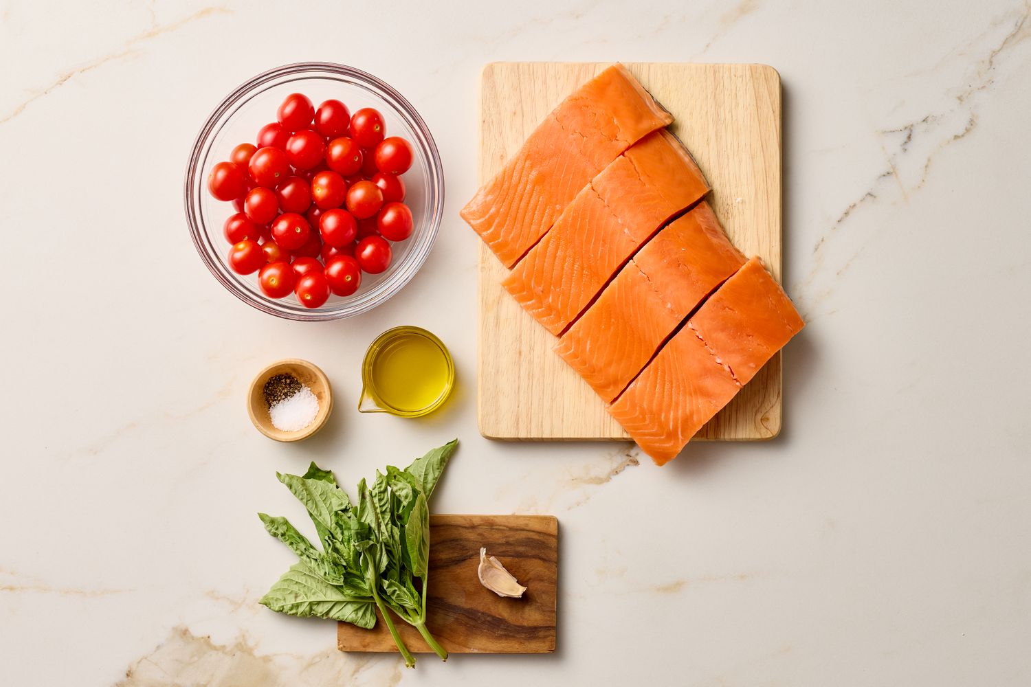 Ingredients consisting of salmon fillets cherry tomatoes olive oil basil leaves garlic and a small bowl of seasoning on a marble countertop