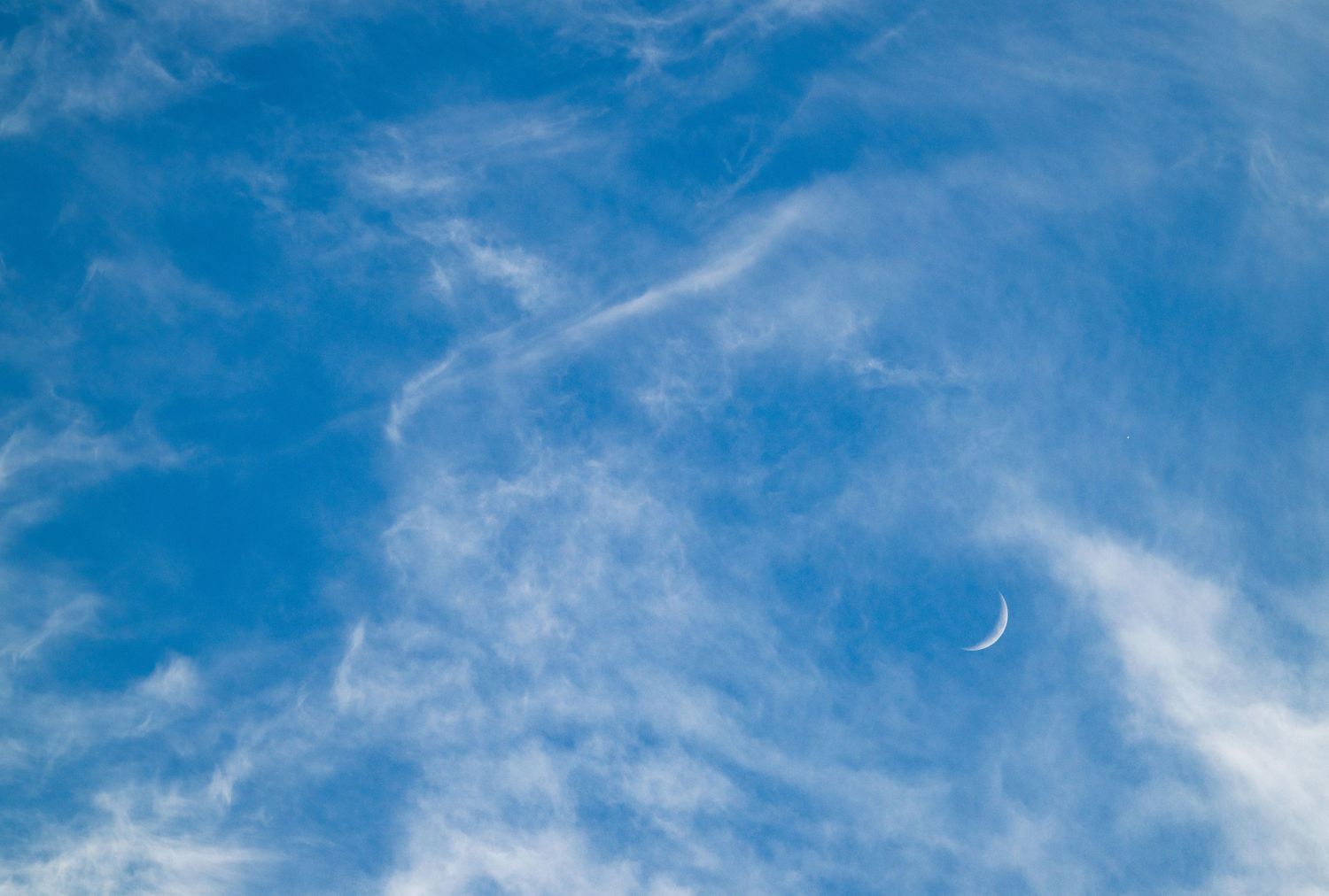 A crescent moon visible in a daytime sky scattered with clouds