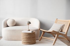 A minimalist living space with a sofa a rounded rattan side table a small lamp and a wooden chair with a cushion