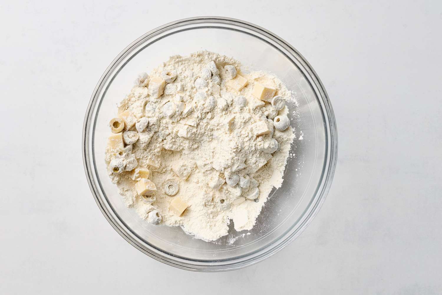 A glass bowl with dry ingredients being prepared for a loaf featuring chunks of cheese and sliced olives visible among the flour