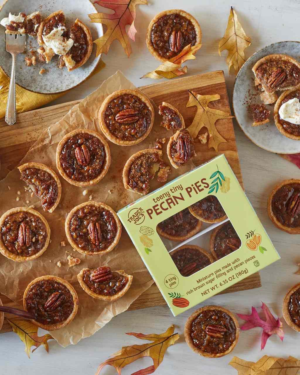 Small pecan pies arranged on a wooden platter with a product box labeled Mini Pecan Pies scattered autumn leaves as decoration