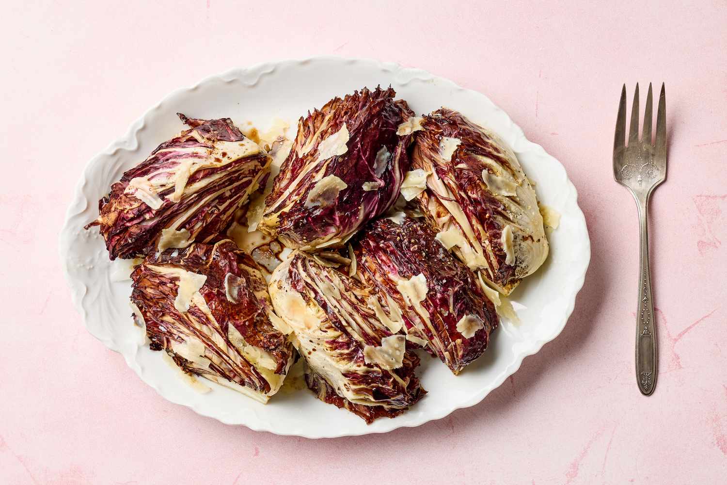 A plate of roasted radicchio wedges with shaved cheese on top displayed alongside a fork on a light background