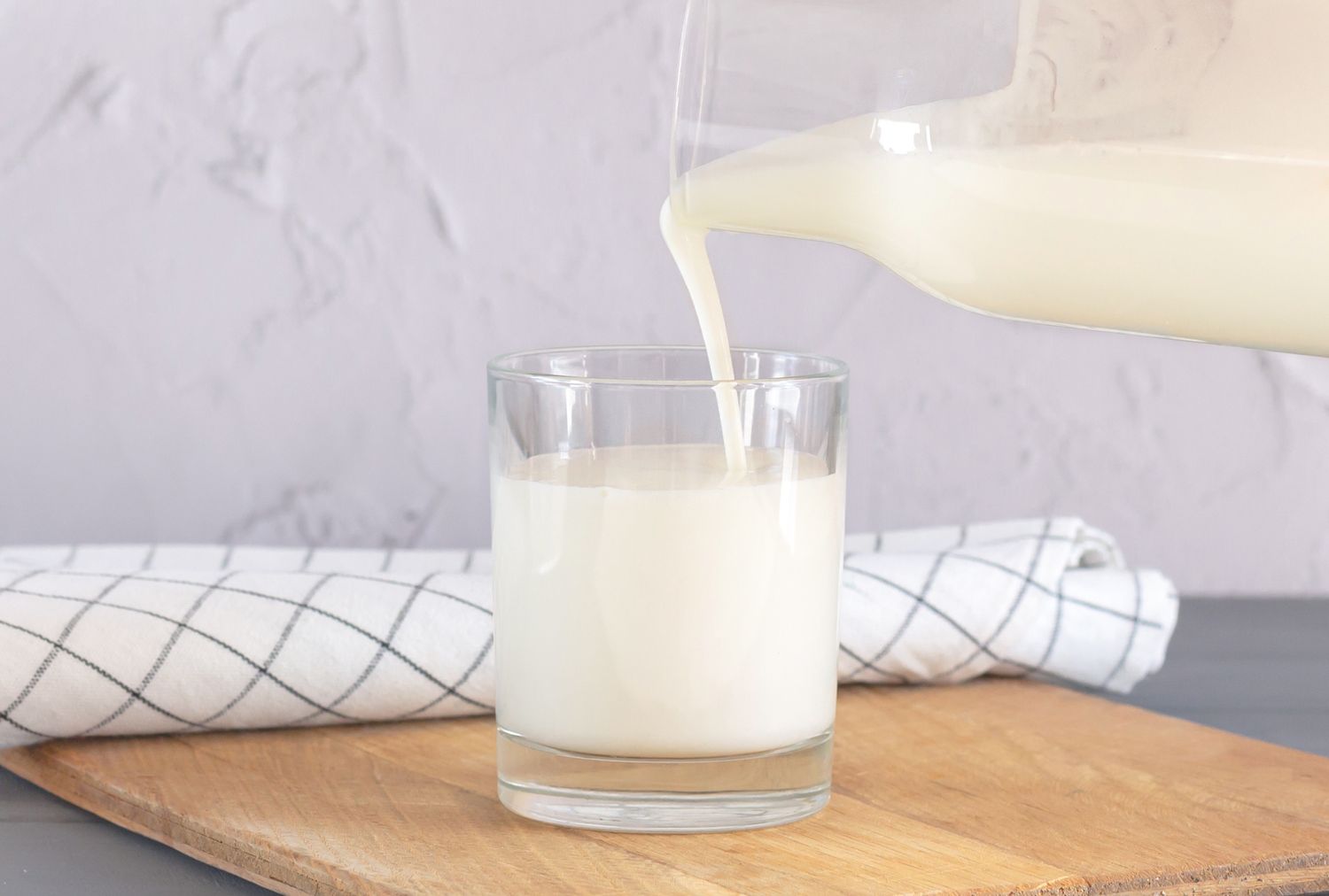 A glass of milk on a wooden surface being poured from a pitcher