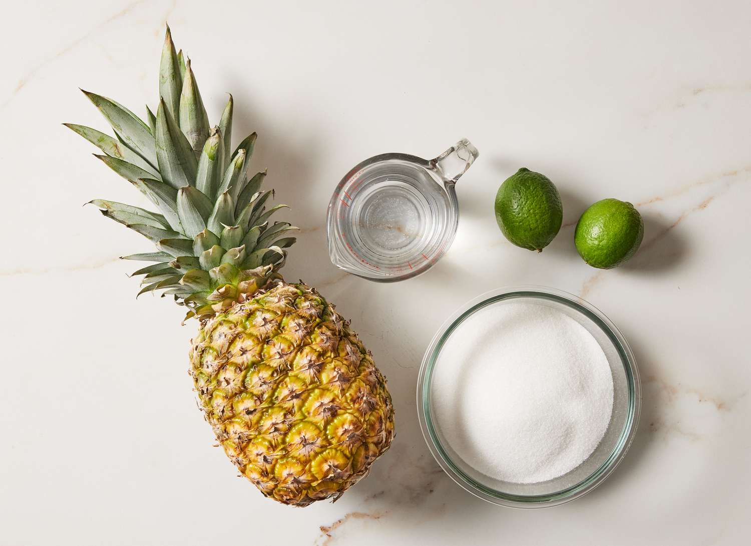 overhead view of full pineapple, limes ad bowl of sugar