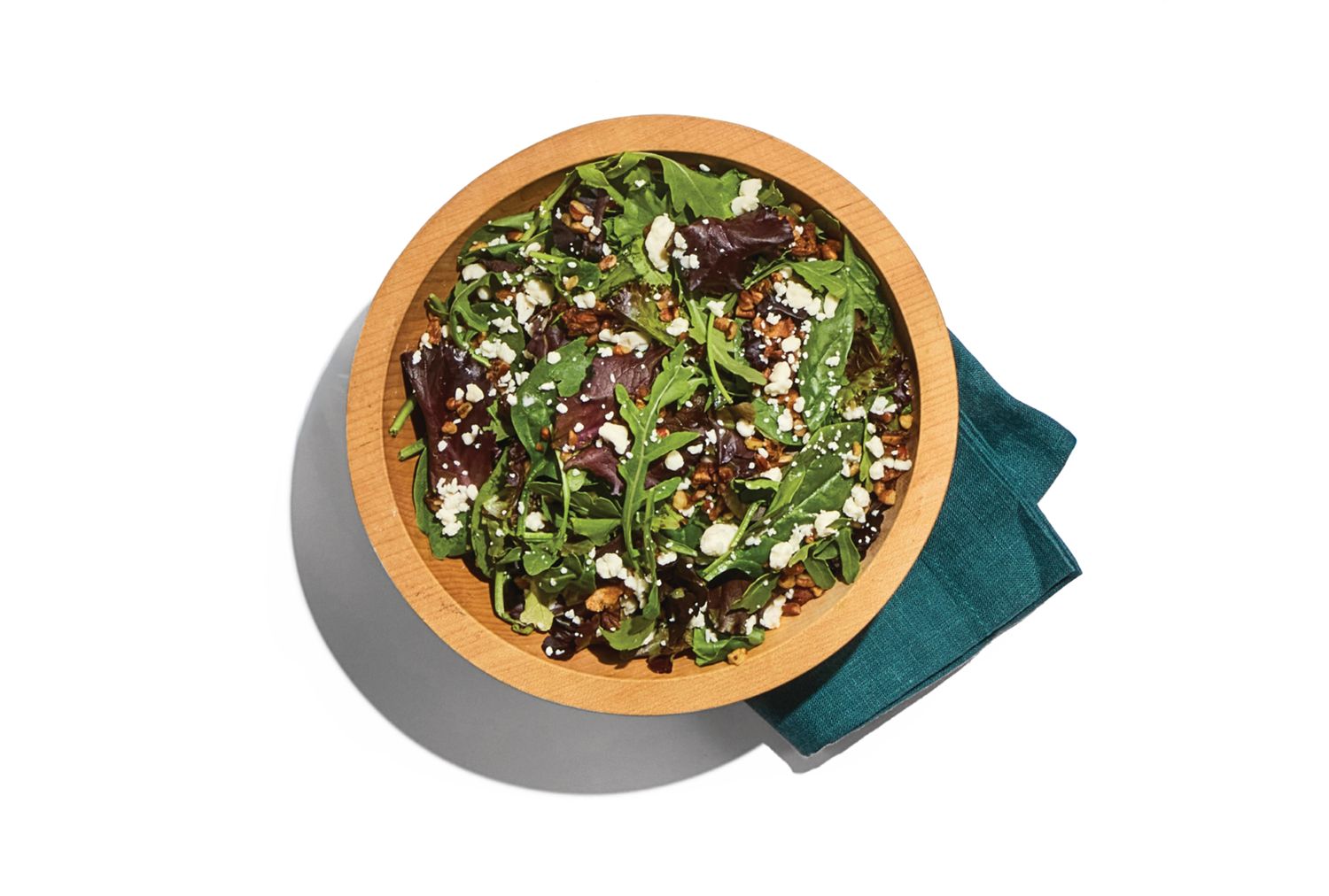 A wooden bowl filled with a leafy green salad including various greens nuts and crumbled cheese placed alongside a folded cloth napkin