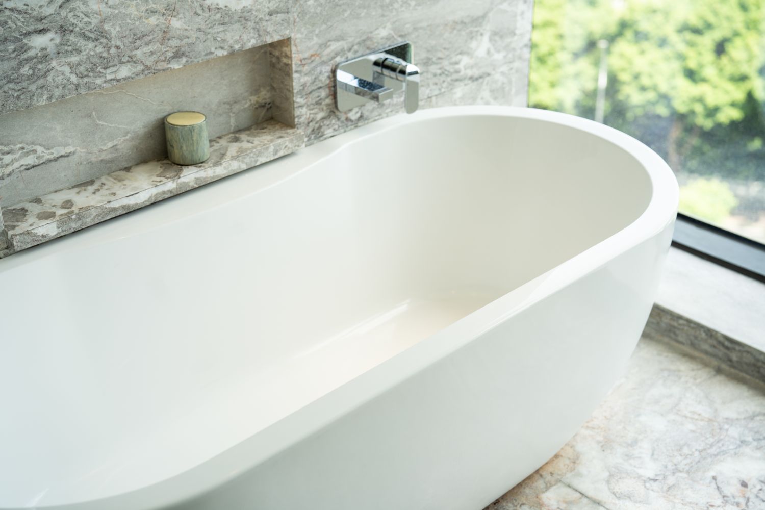 A modern freestanding bathtub with a view of a garden through a nearby window