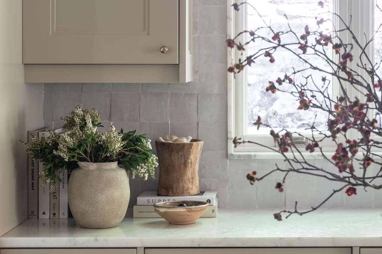 A kitchen counter with a vase of flowers a wooden container books and a branch with blossoms by the window