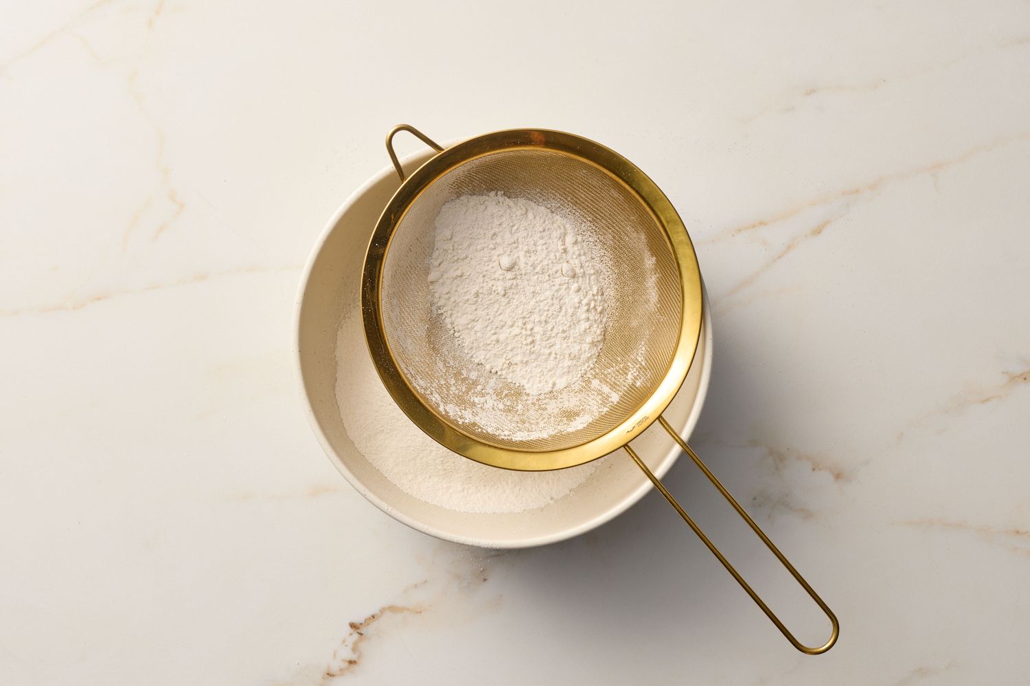 A bowl and sieve with sifted flour on a marble surface, kitchen preparation scene