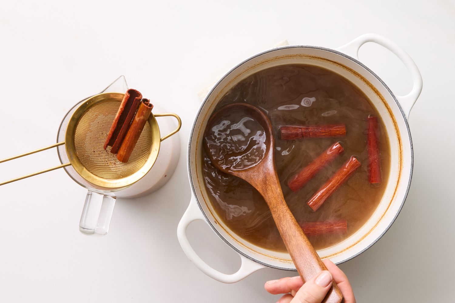 Cooking pot with spiced cider and cinnamon sticks hand stirring with a wooden spoon strainer with cinnamon sticks on the side