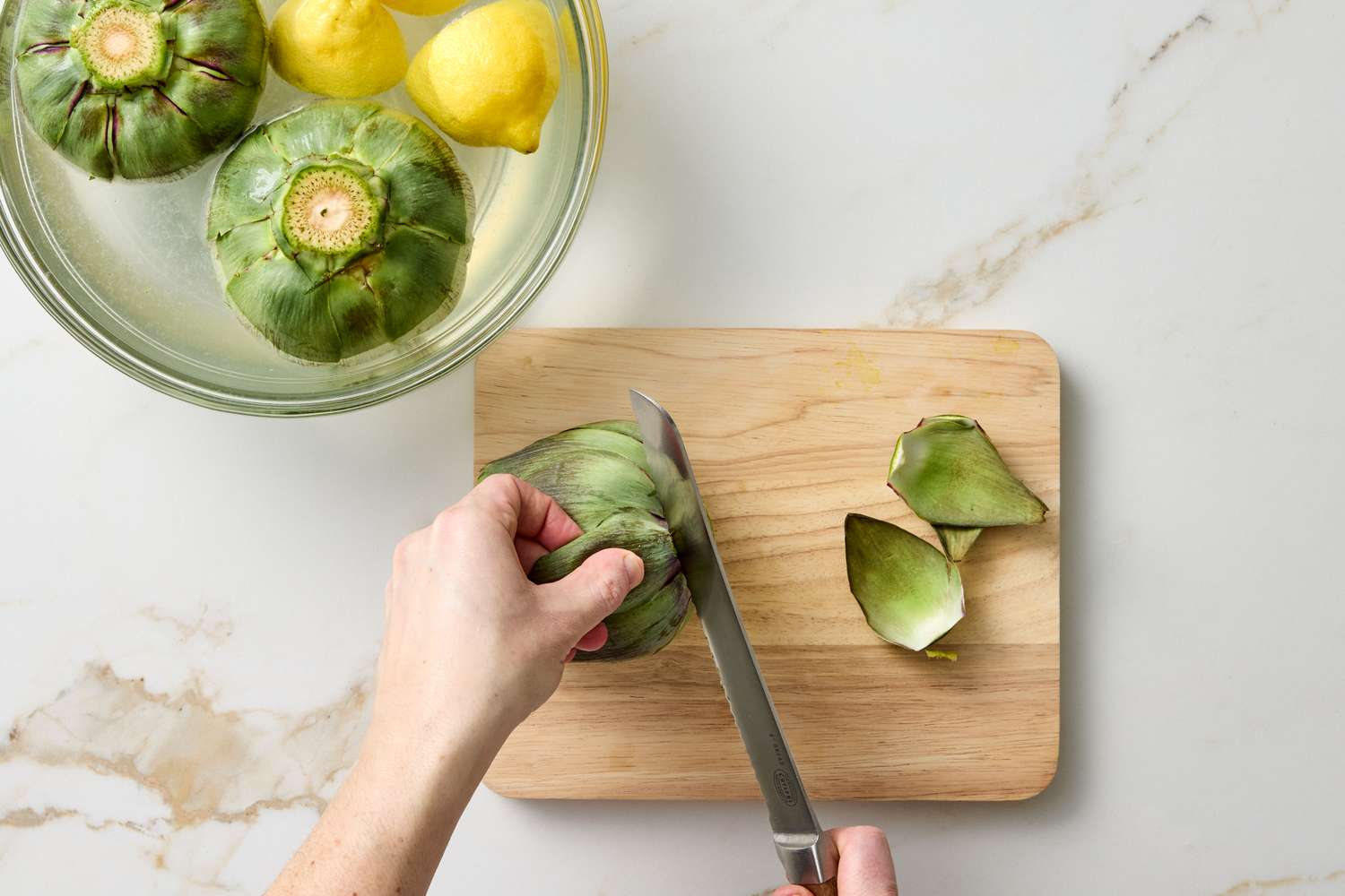 A hand shown cutting an artichoke on a wooden cutting board with a bowl containing whole artichokes and lemons in the background