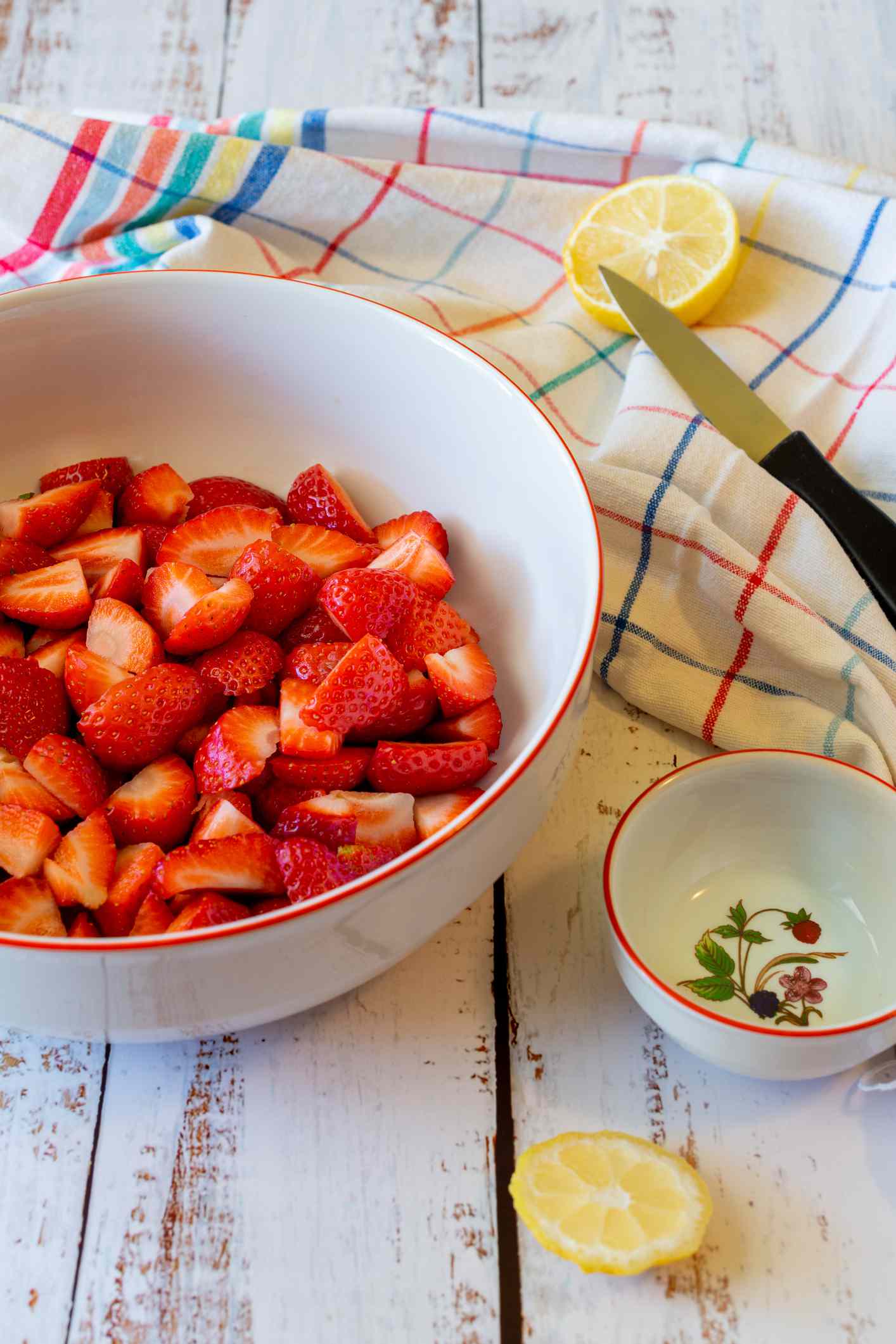 A bowl of chopped strawberries near a lemon wedge, a knife, and a decorative bowl