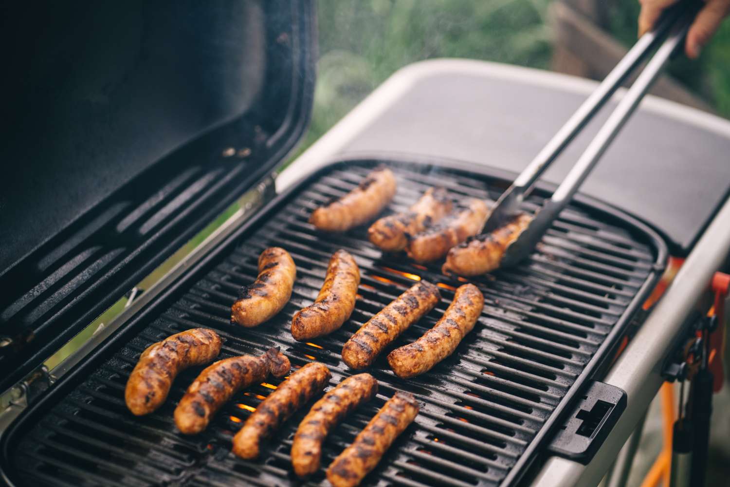 Grilling sausages on a barbecue grill with a hand using tongs to turn them