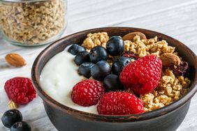 granola with greek yogurt, nuts and fresh berries in a bowl with cereal in jar over white wooden background