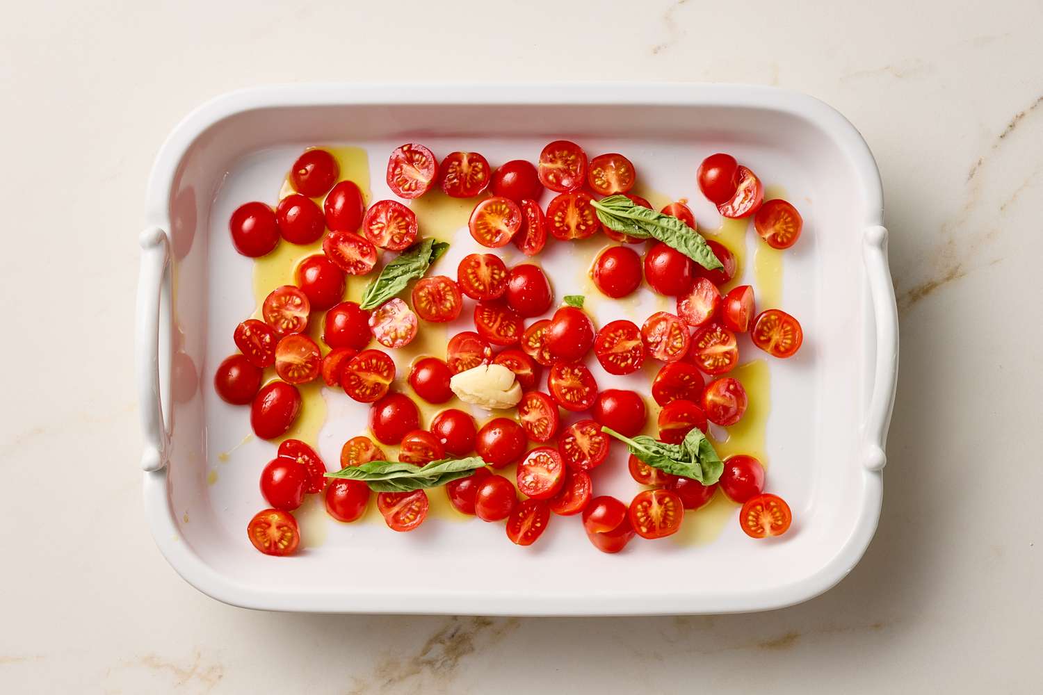 A baking dish with halved cherry tomatoes basil leaves and a clove of garlic drizzled with oil