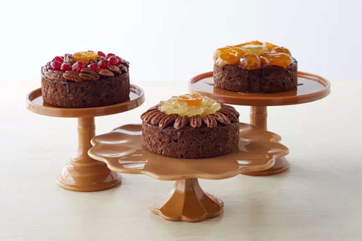 Three fruitcakes presented on wooden cake stands each topped with different fruit arrangements